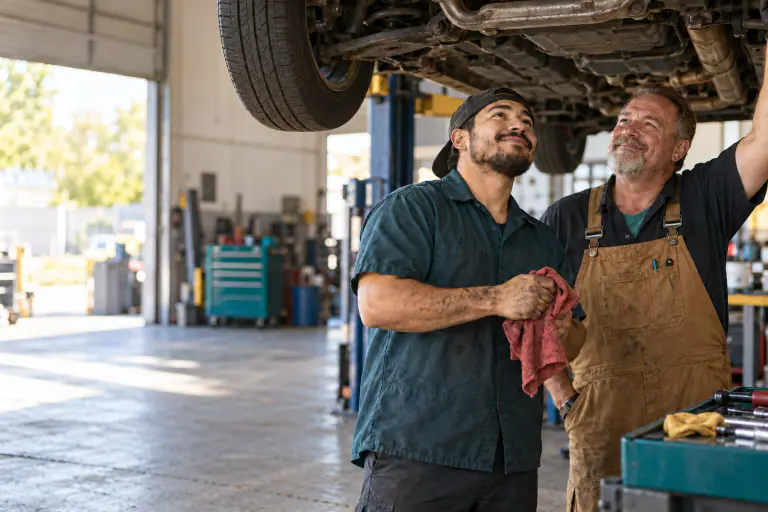Mechanic and shop owner under a lifted car, wiping hands on a red rag