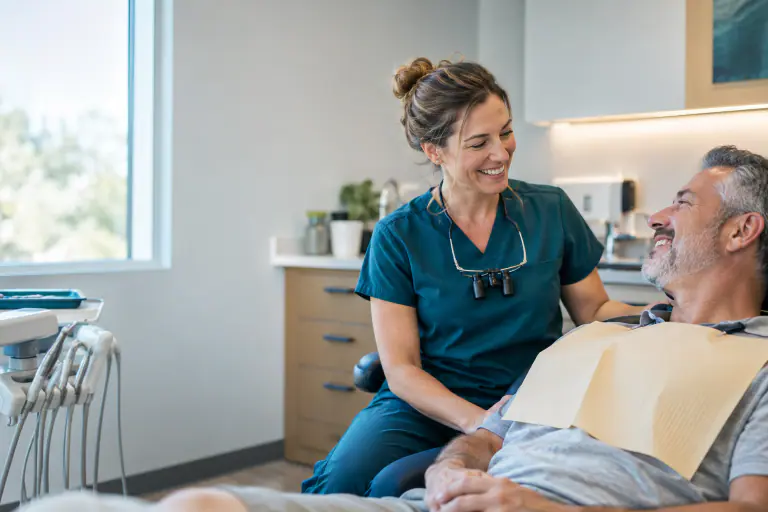 Dentist in scrubs and loupes smiling at a patient in a modern exam room