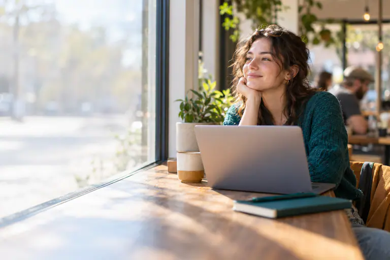 Freelance writer in her 20s working on a laptop at a sunny cafe window