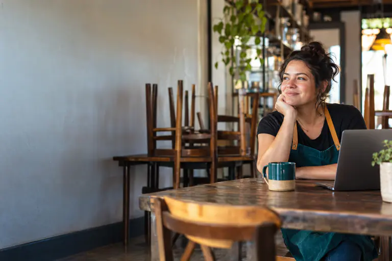 Small restaurant owner in an apron sitting at a back table after close, laptop open, chairs stacked behind her